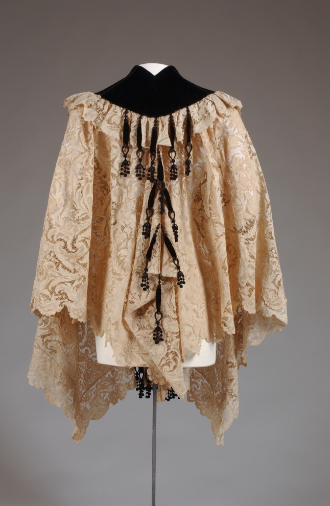 The House of Worth, Cape, Circa 1890. Paris. Gift of the Estate of Elizabeth Arden. 69.160.9 Hip-length cape in cream lace with wide neckline border of black silk velvet; trimmed with black silk chenille bobble tassels